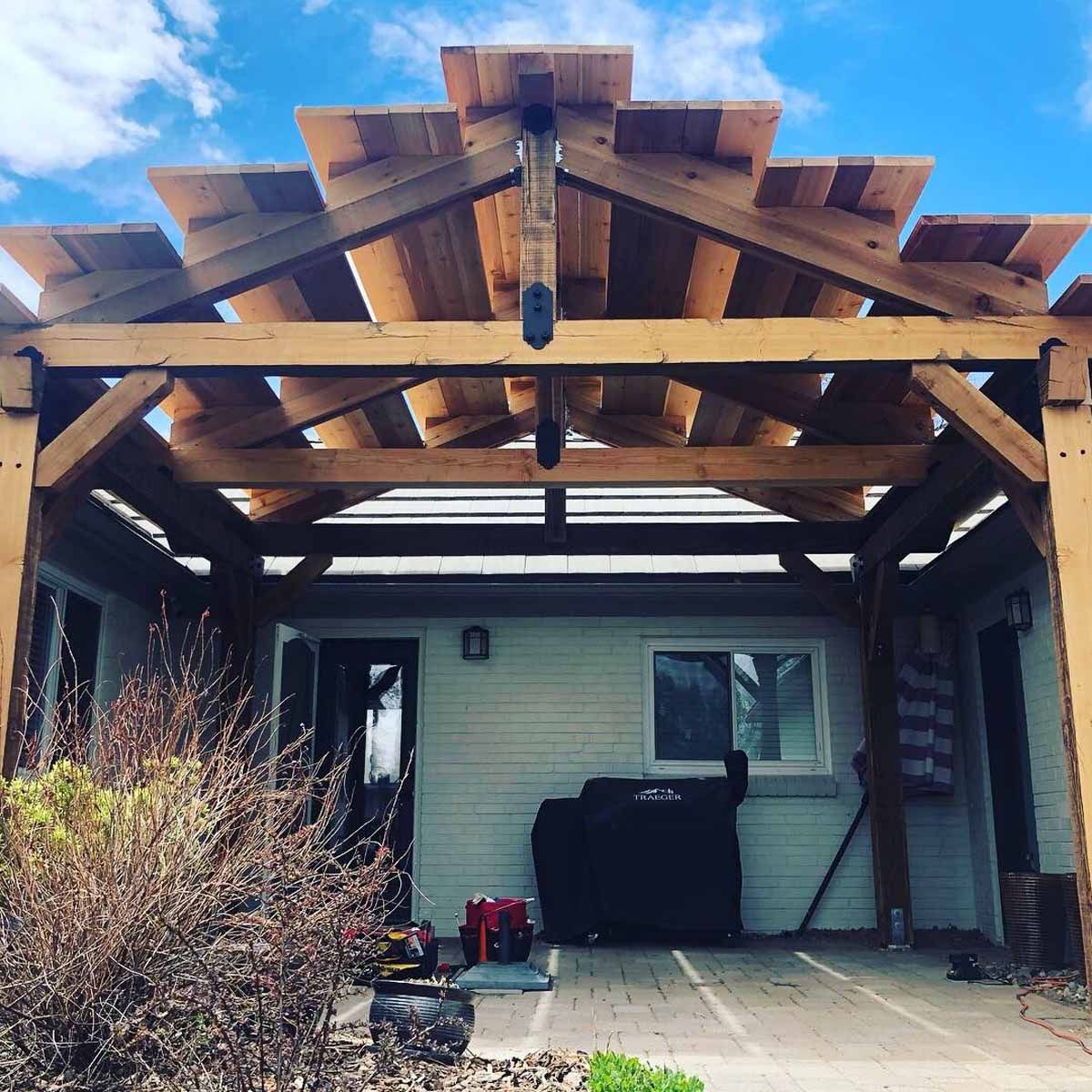 Back patio with unfinished wooden pergola framing a house wall, grill, and potted plants under a blue sky