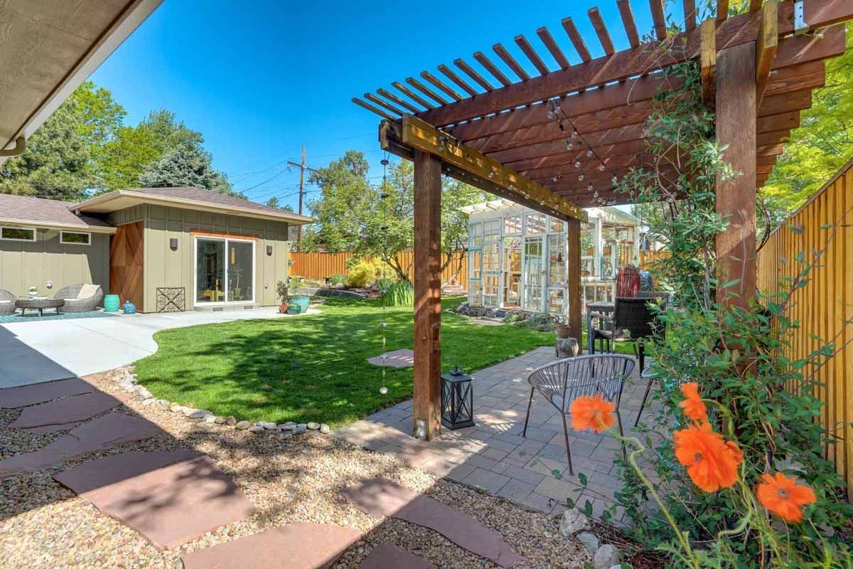 Backyard patio with wooden pergola, orange flowers, green lawn, and a small shed under a bright blue sky