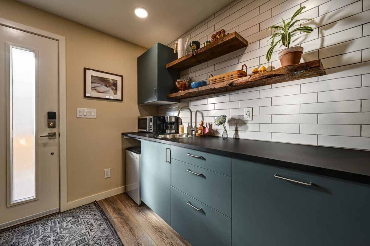 Narrow mudroom with teal cabinets, black countertop, white subway tile, and wood shelves by a glass door