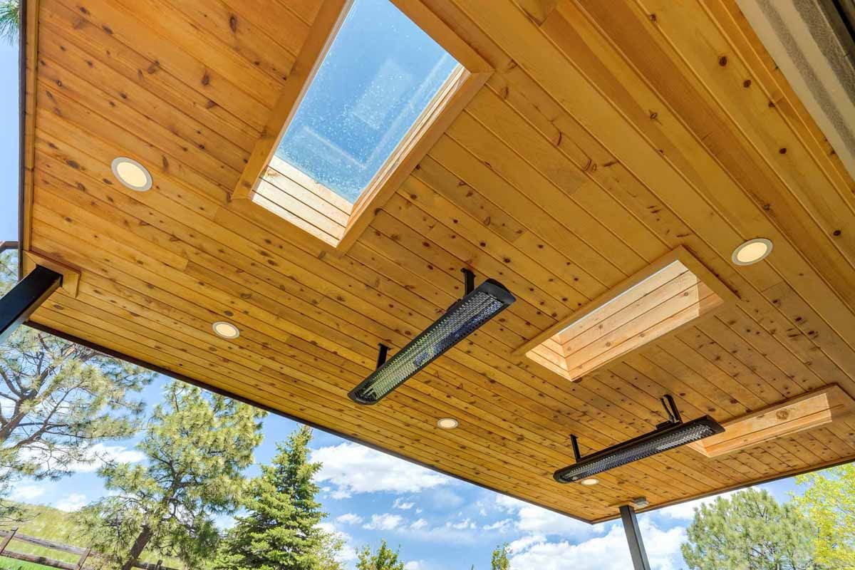 Wooden patio ceiling with skylights and recessed lights, viewed upward against a blue sky and trees
