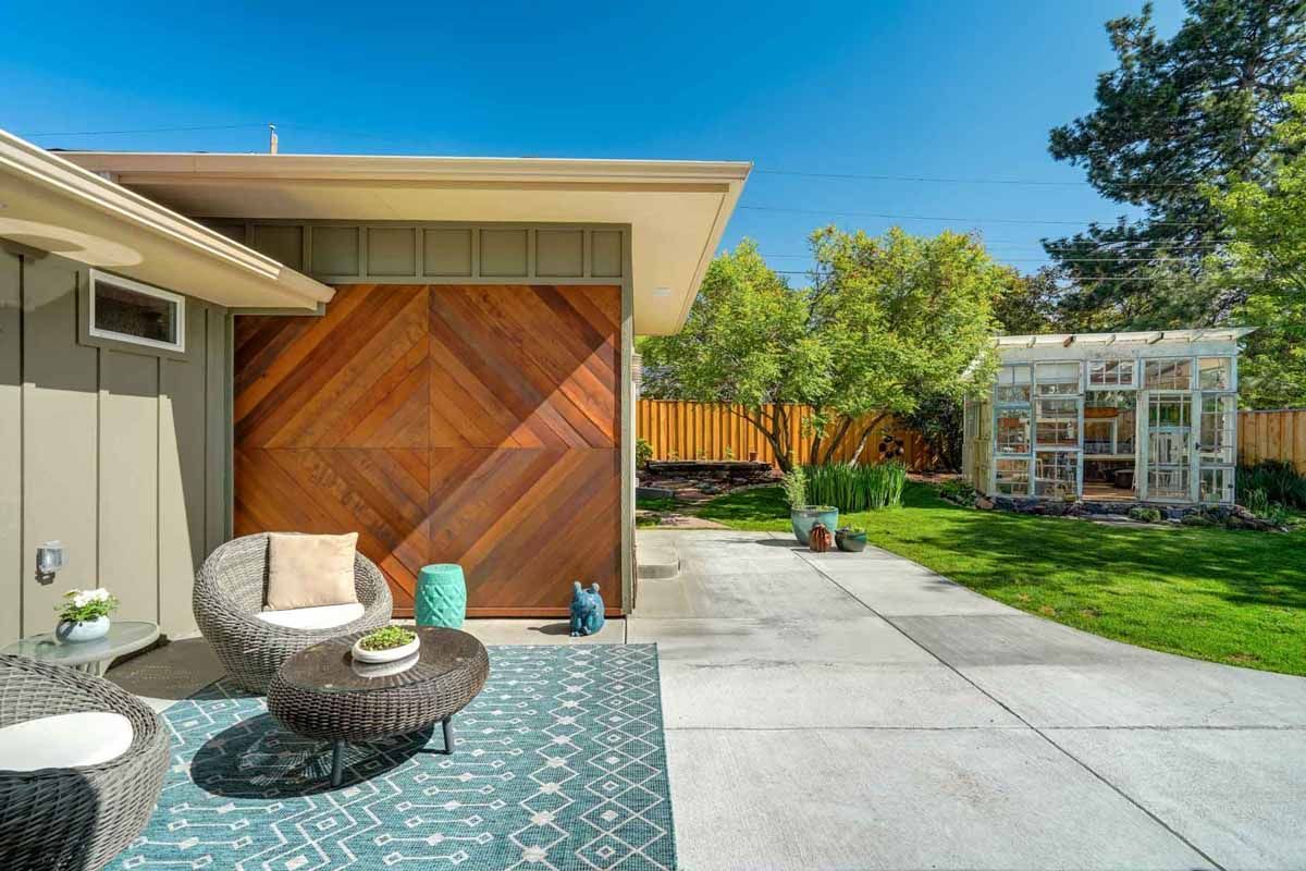 Patio with wicker chairs, patterned rug, wood accent wall, and a concrete path beside a bright backyard garden
