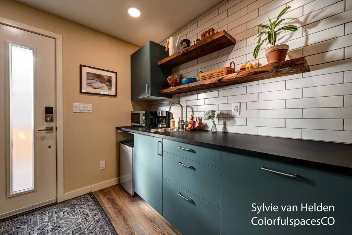 Modern mudroom with teal cabinets, black countertop, white subway tile, wood shelves, and a glass door