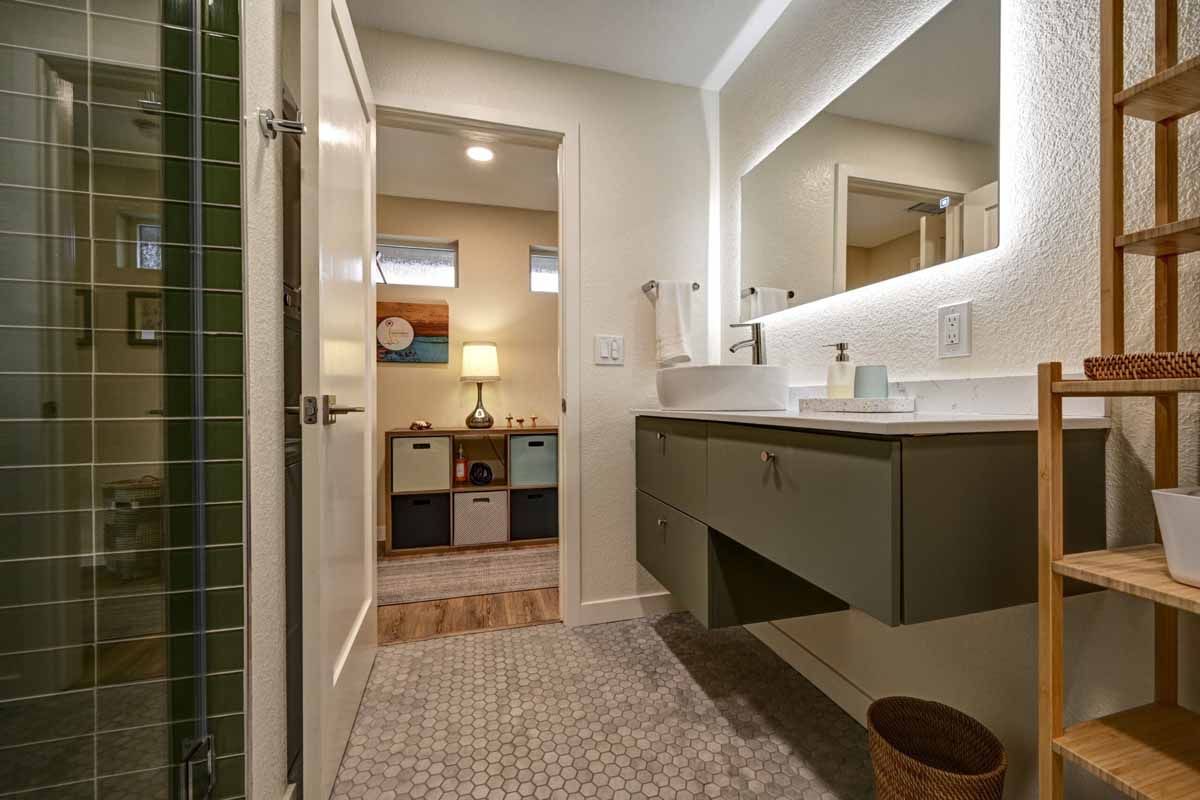 Modern bathroom with green tile shower, long vanity, large mirror, and bedroom visible through doorway