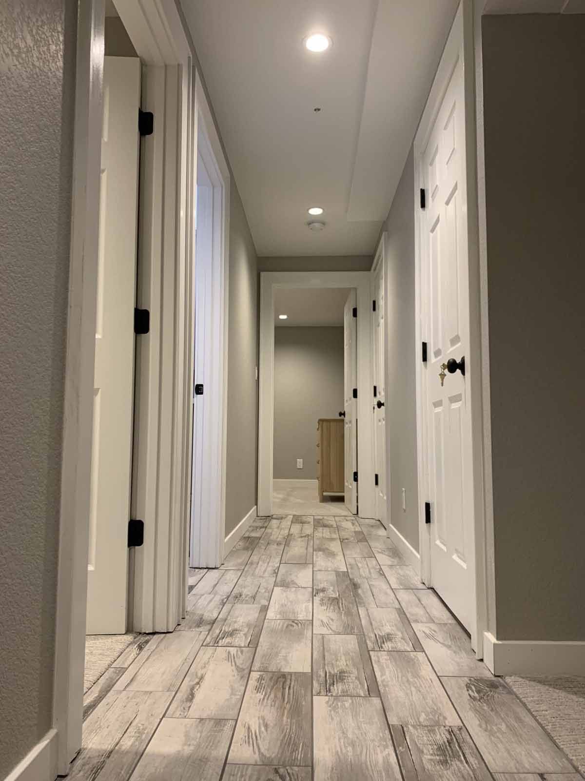 Narrow hallway with gray wood-look floors, white walls, recessed lights, and several doorways.