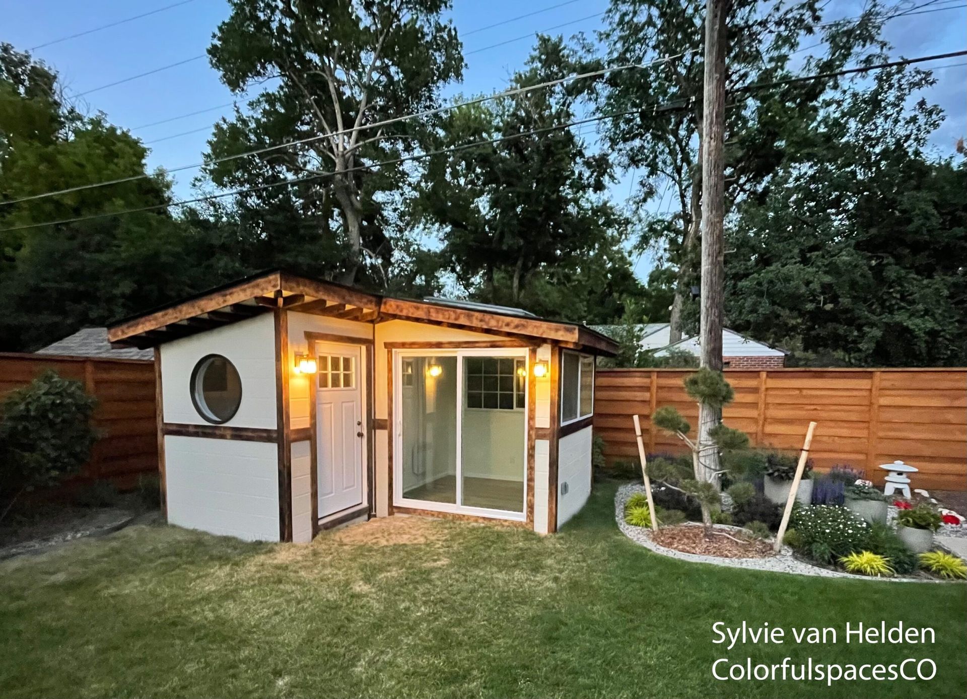 Small white garden shed with warm lights, glass doors, and wood fencing in a grassy backyard at dusk