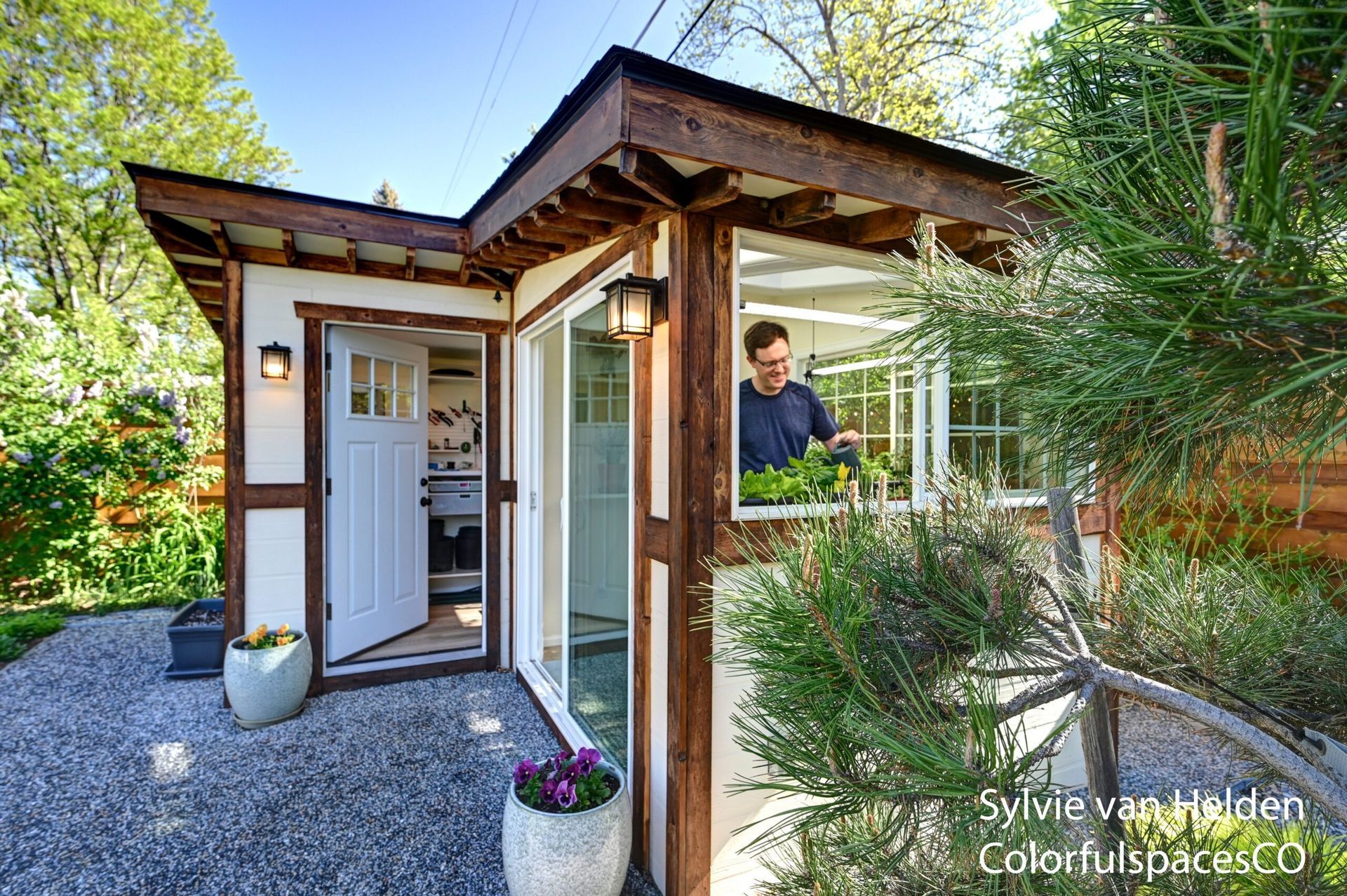Front entrance of a modern home with wood trim, gravel path, potted flowers, and a person standing inside the doorway