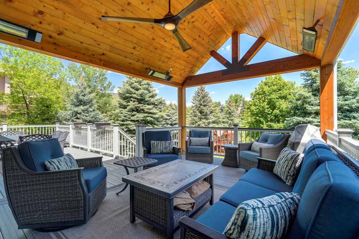 Covered outdoor patio with wicker seating, blue cushions, and a wooden vaulted ceiling overlooking trees.