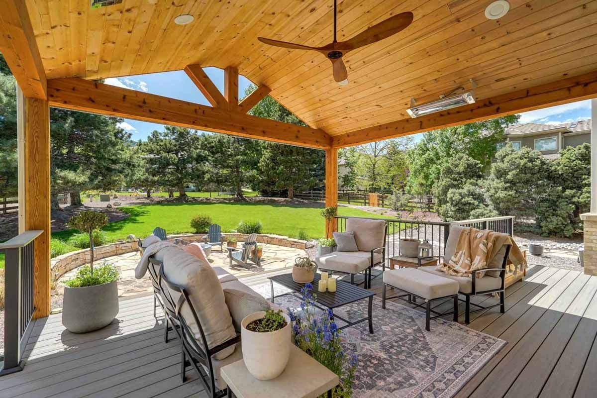 Covered wooden patio with outdoor seating, potted plants, and a view of a green backyard