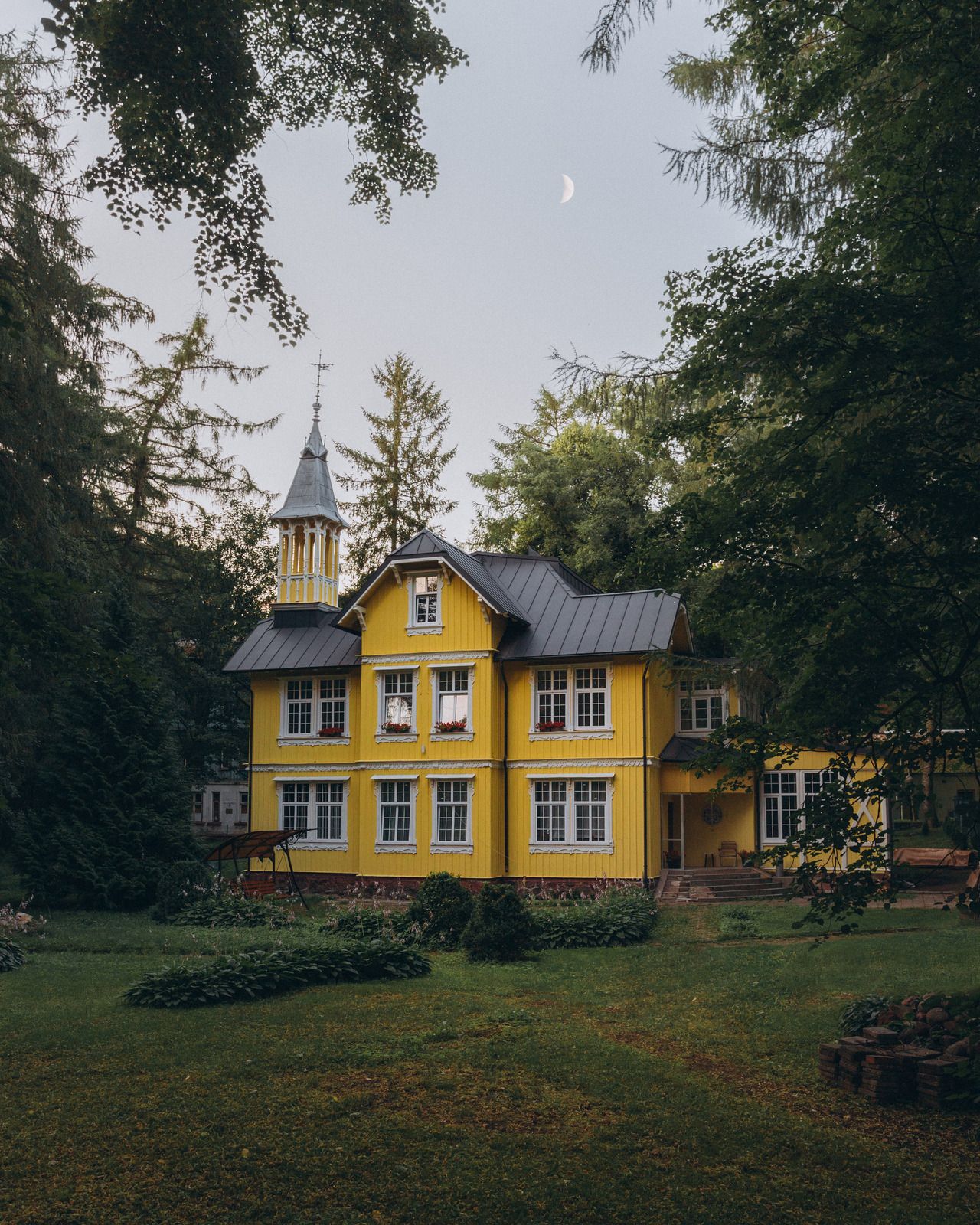 Yellow two-story house with a tower and dark roof, surrounded by trees. A crescent moon is visible in the sky.