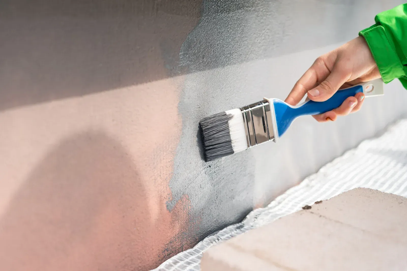 Person painting a wall with a blue-handled brush; hand and green sleeve visible.