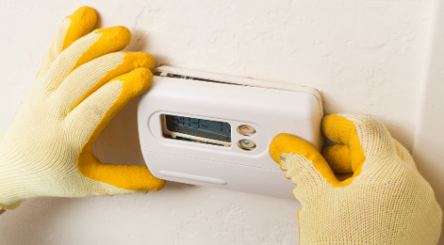 A person wearing yellow gloves is installing a thermostat on a wall.