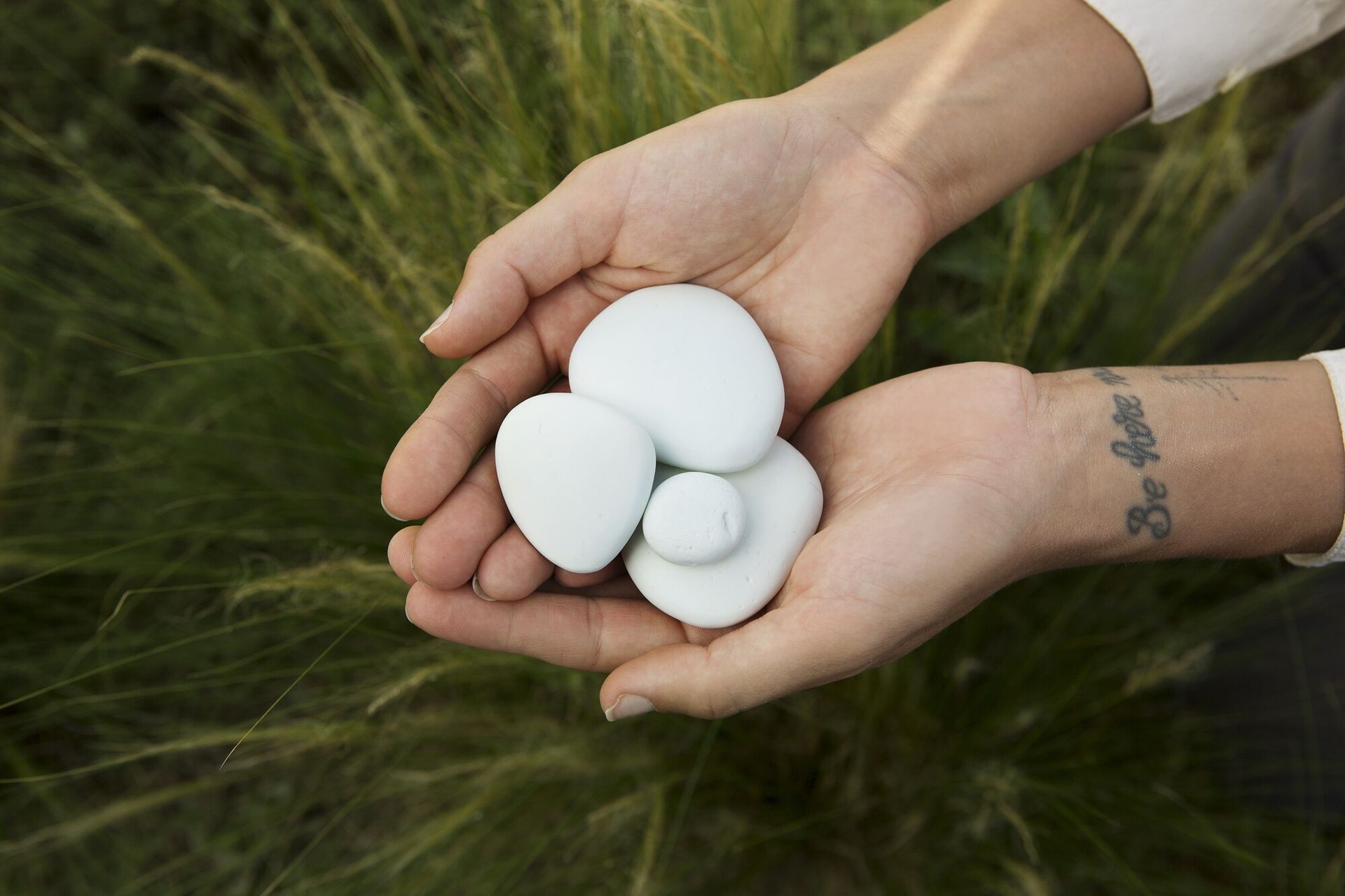 Hands cupping four smooth, white stones against a background of green grass. A tattoo is visible on the wrist.