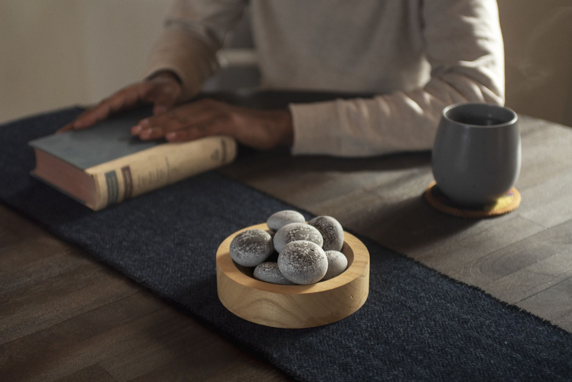 Person at table with book, cup, and wooden bowl of treats on a dark blue runner.