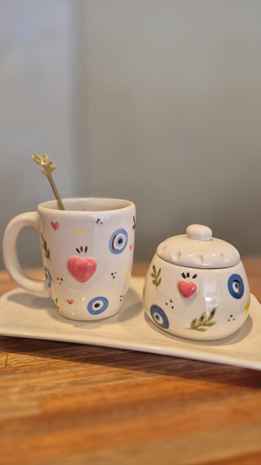 Taza pequeña de cerámica y azucarero con coloridos dibujos de corazones y ojos sobre una mesa de madera.