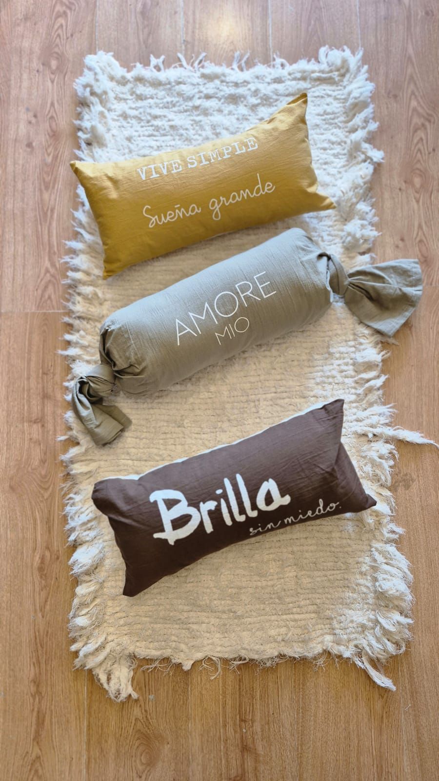 Tres pequeños cojines decorativos bordados sobre una alfombra blanca de pelo largo, en tonos dorados, grises y marrones.