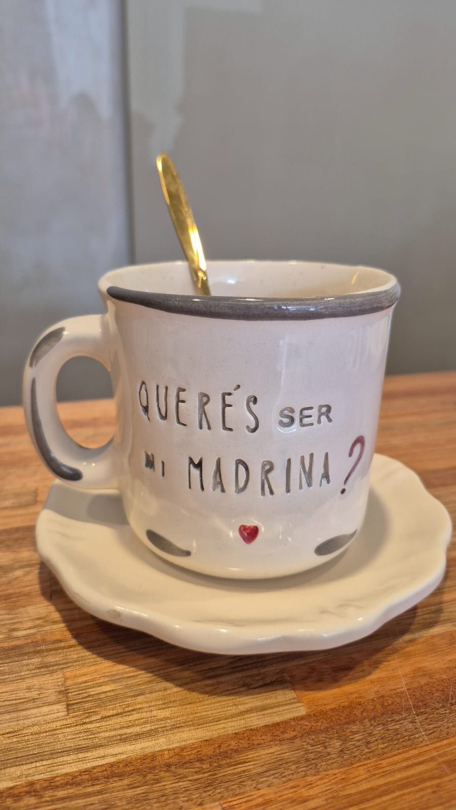 Taza de cerámica blanca sobre platillo con una cuchara dorada, que dice 