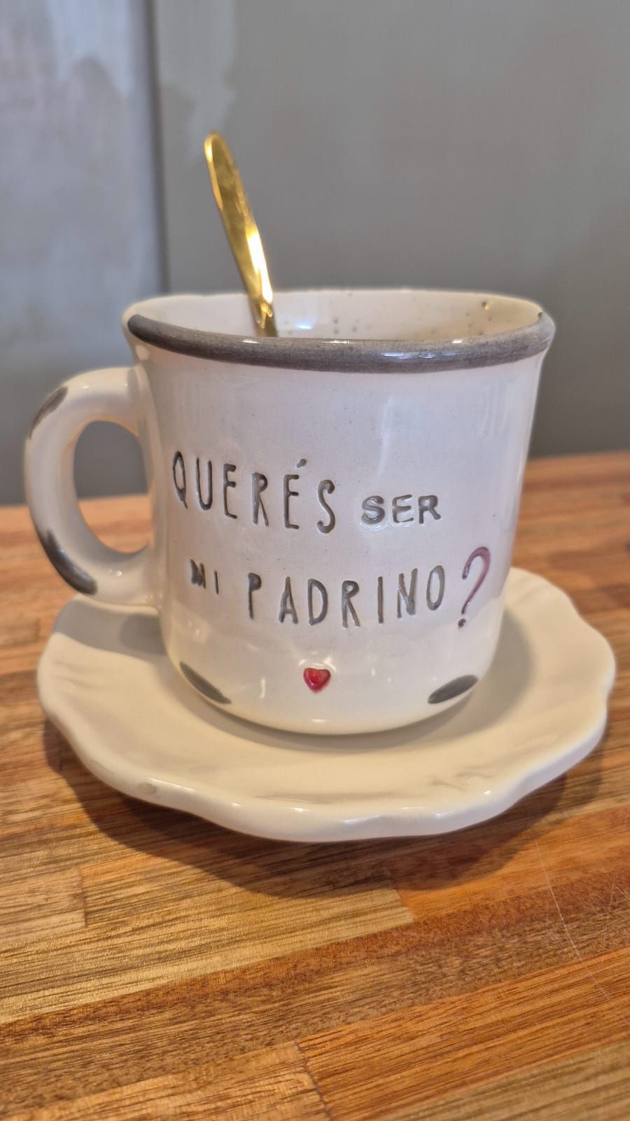 Taza de cerámica blanca sobre platillo con una cuchara dorada, que dice 