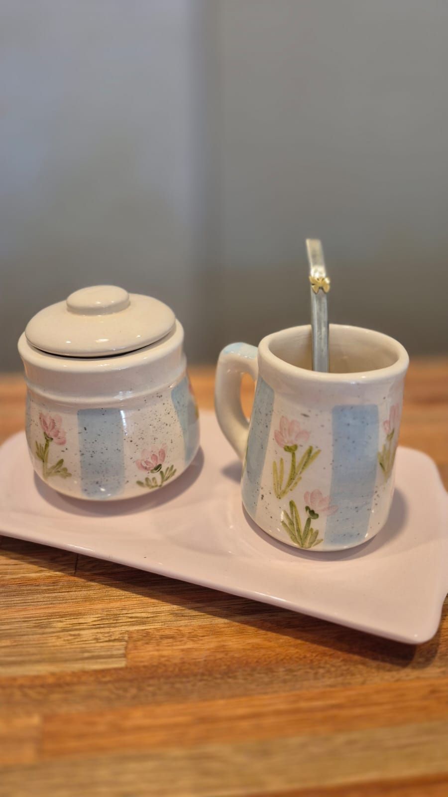 Azucarero de cerámica, taza con cuchara y bandeja sobre una mesa de madera.