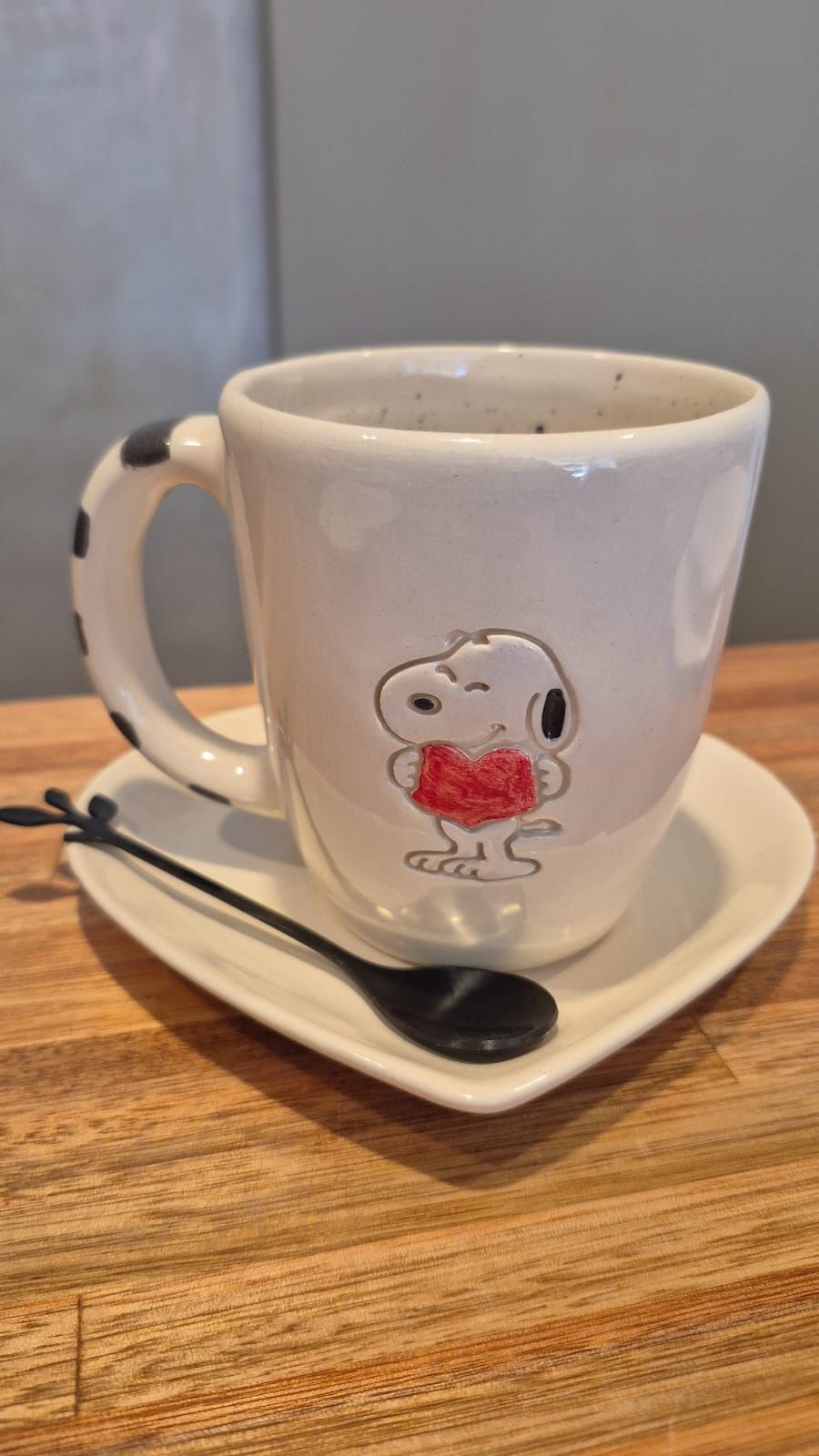 Taza blanca con un diseño de Snoopy en un platillo, con una cuchara, sobre una mesa de madera.
