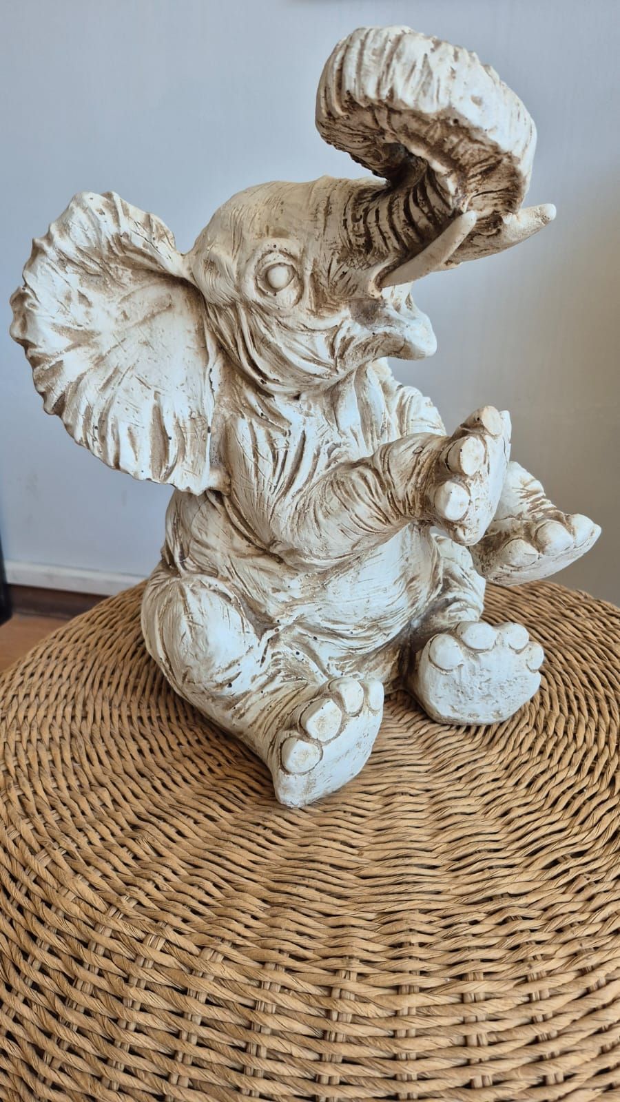 Figura de elefante blanco con trompa levantada sobre una mesa redonda tejida contra una pared beige.