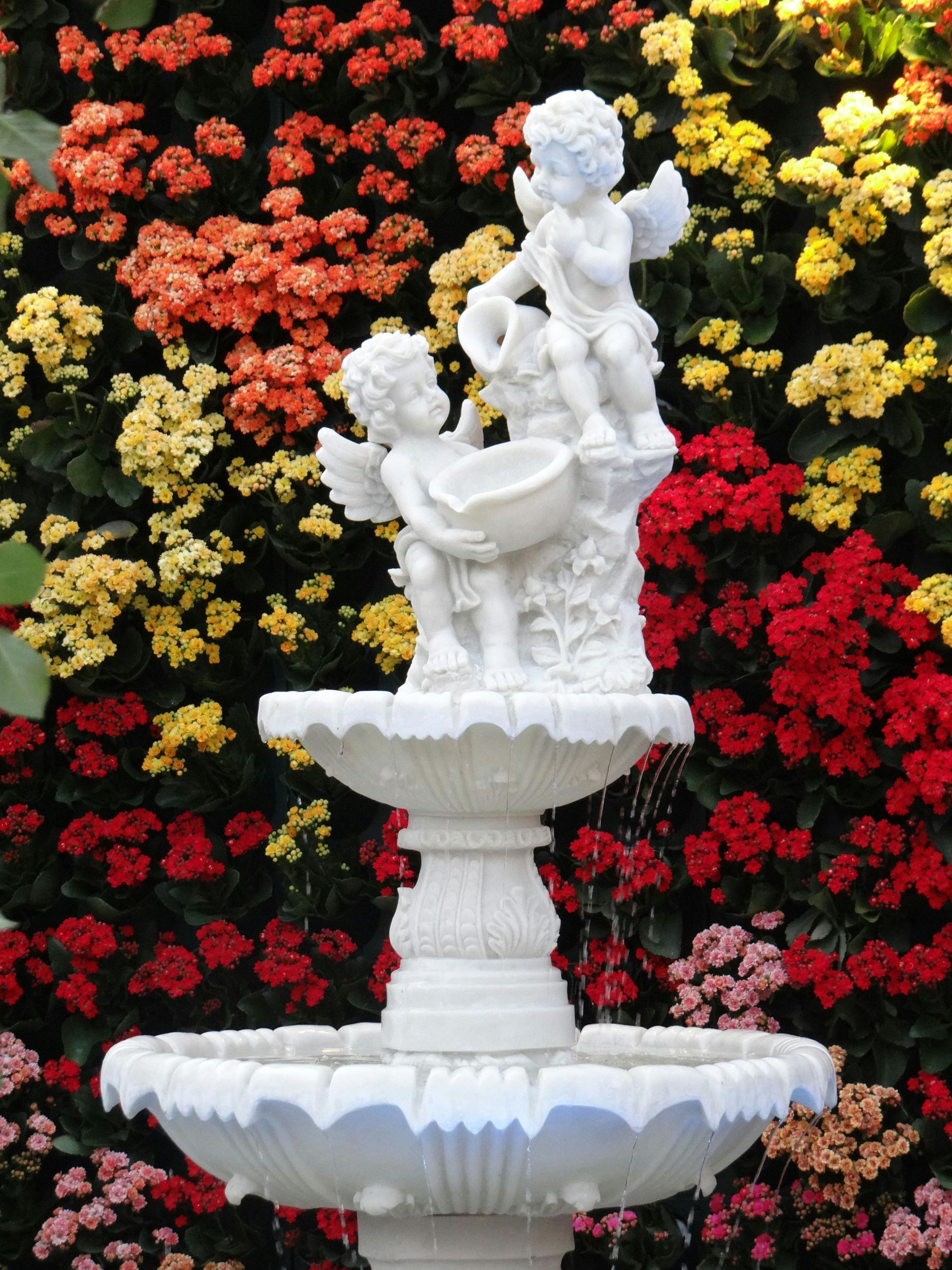 Estatua de fuente blanca sobre un fondo denso de flores rojas, amarillas, rosas y negras.