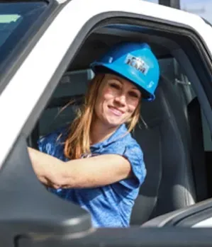 Woman in a blue hard hat smiles while driving a white truck.