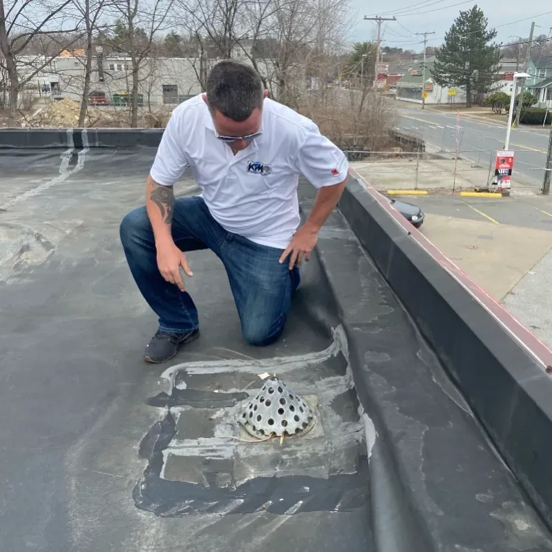 Man kneeling on a black rooftop, inspecting a drain. He wears a white shirt and jeans.
