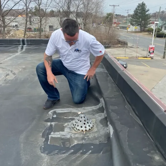 Man kneeling on a black rooftop, inspecting a drain. He wears a white shirt and jeans.