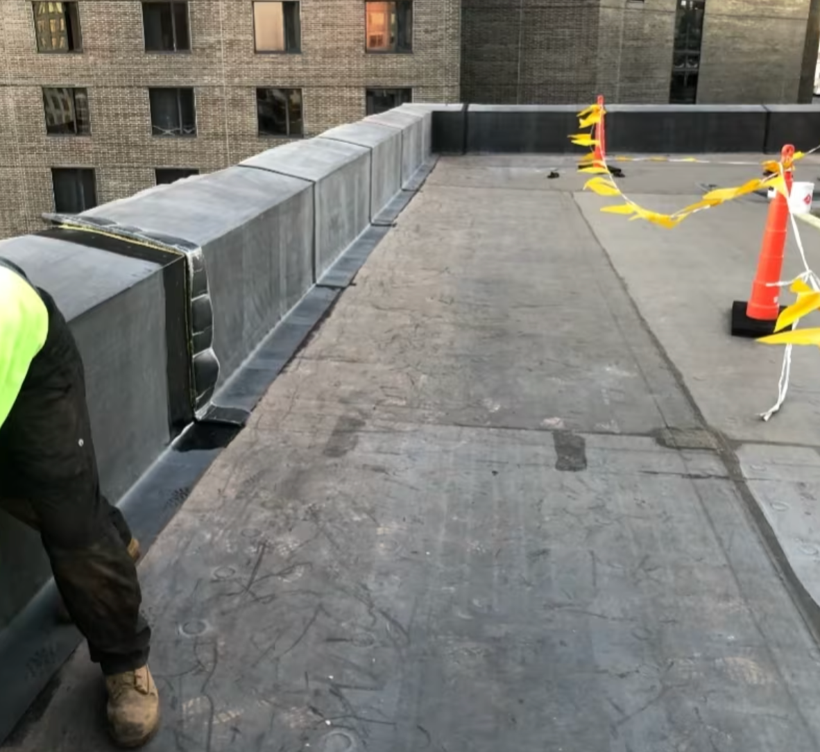Person working on a flat, black roof. Yellow caution tape and orange cones mark a perimeter. Buildings in background.