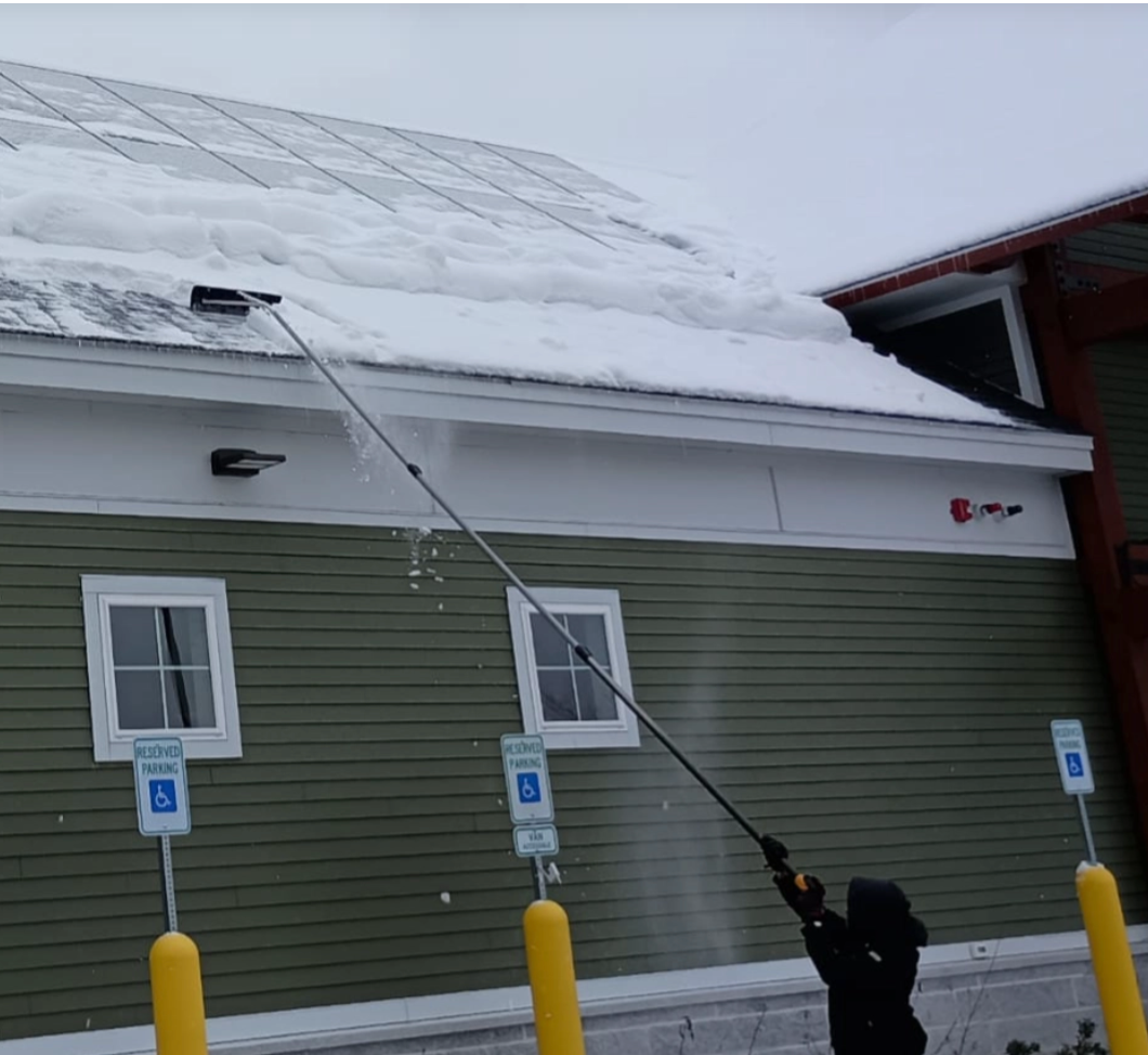 Person using a long-handled snow removal tool to clear snow from a building's rooftop solar panels.