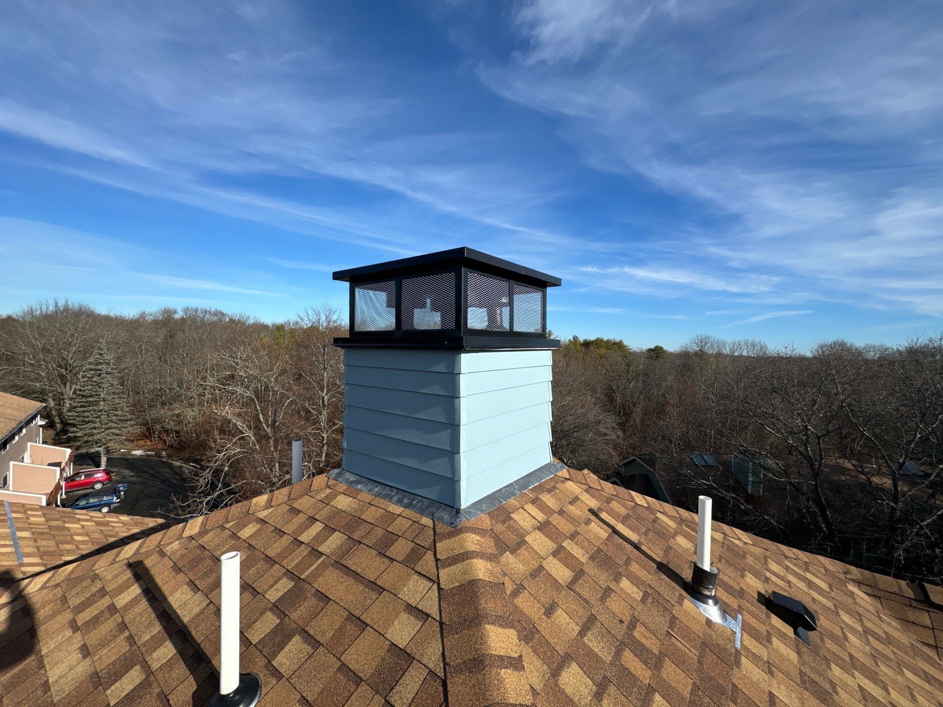 Chimney on a brown shingled roof, light blue siding, black cap, against a blue sky with trees.