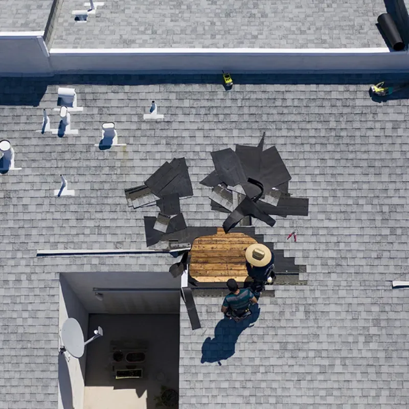 Aerial view of roof repair: Two workers on a gray shingled roof, removing damaged materials.