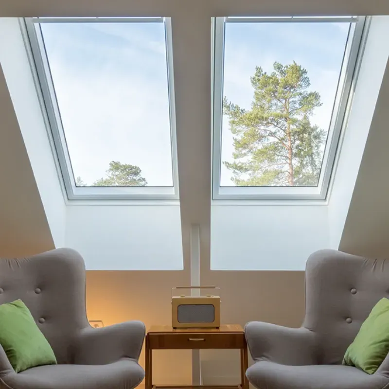Two gray armchairs face small wooden table. Skylights frame tree outside.