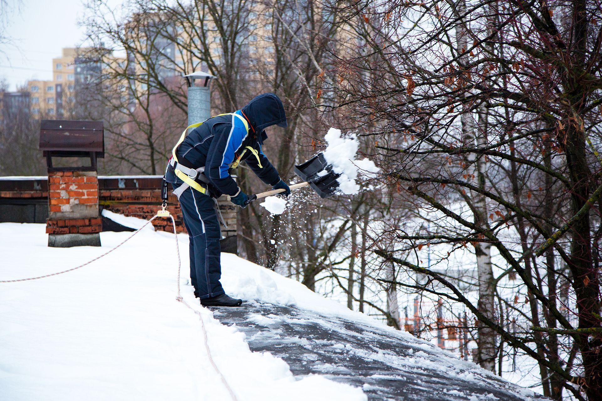 Person shoveling snow off a rooftop, wearing safety harness and winter clothing.