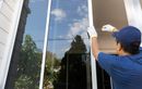 Person in blue shirt and gloves installing a window, outdoors, with a bright sky reflected.