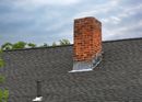 Brick chimney on a dark gray shingled roof under a cloudy sky.