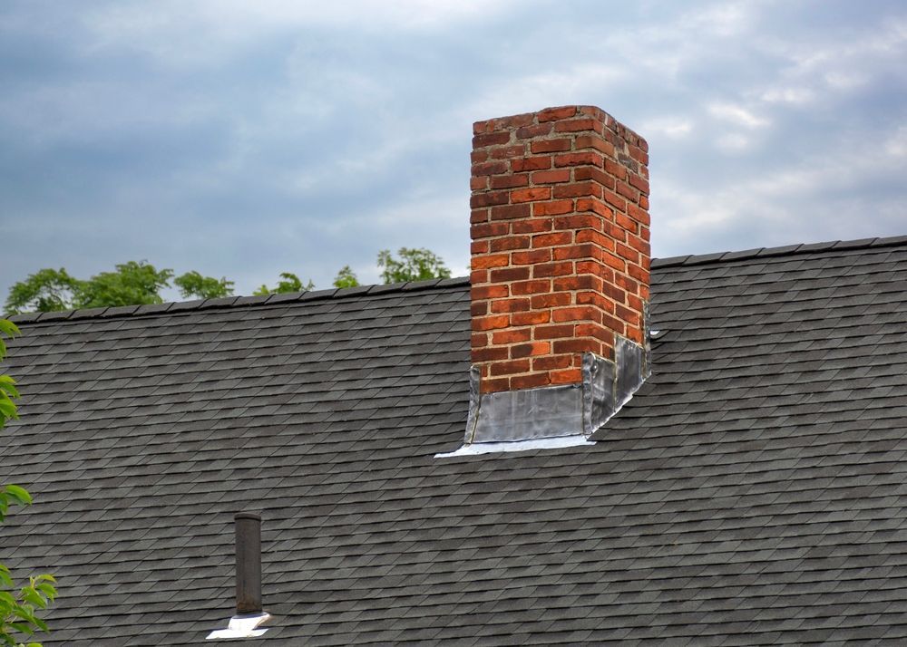 Brick chimney on a dark gray shingled roof under a cloudy sky.