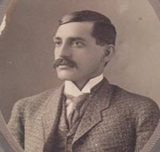 A man in a tweed suit with a mustache, looking off-center.