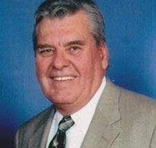 Man in light gray suit, smiling, with a gray and black tie. Blue backdrop.