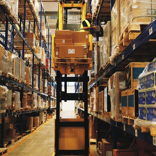 Un hombre está parado sobre una carretilla elevadora en un almacén lleno de cajas.