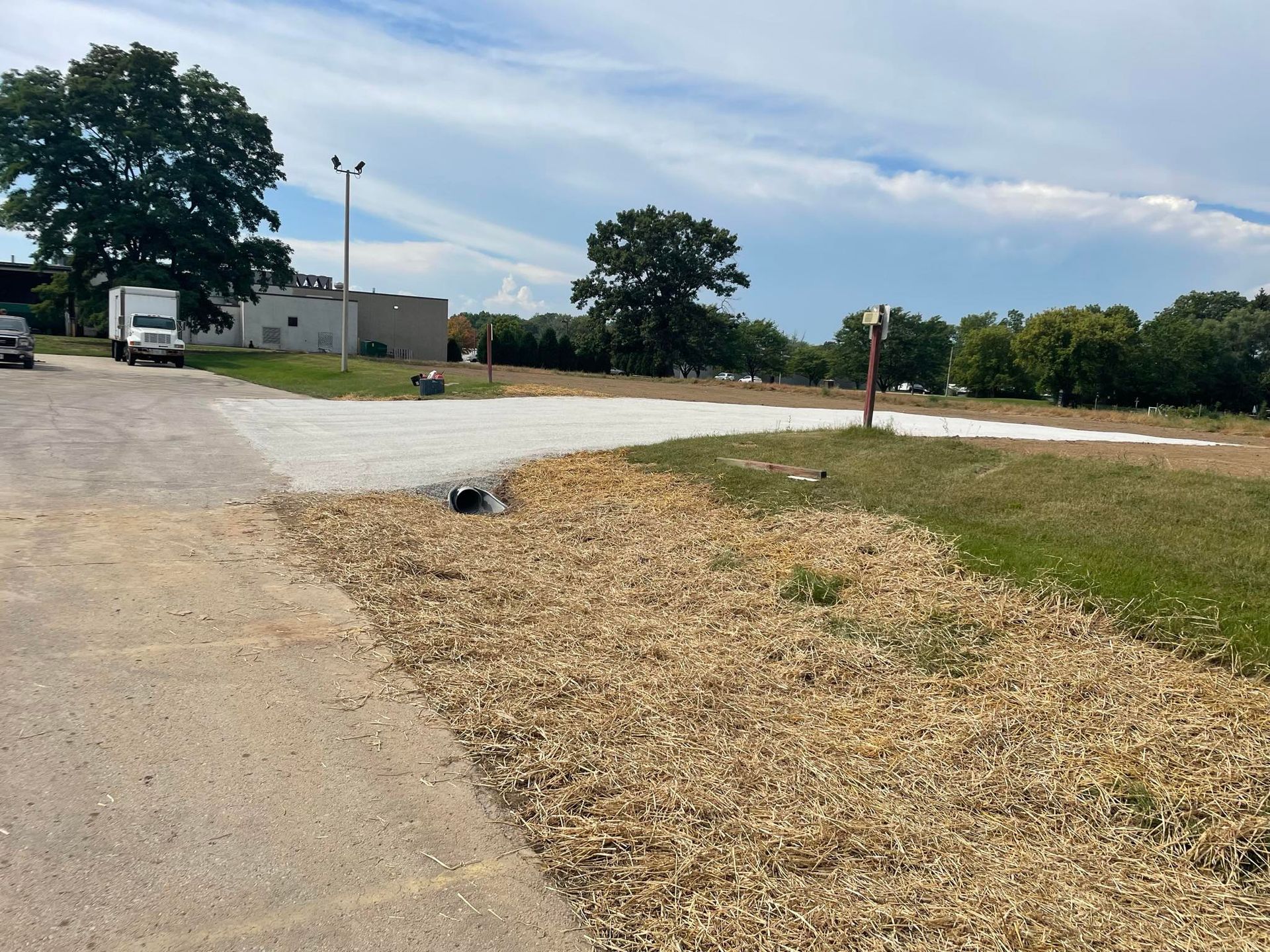 Gravel lot with dry grass border next to a paved road, trees in the background, cloudy sky.