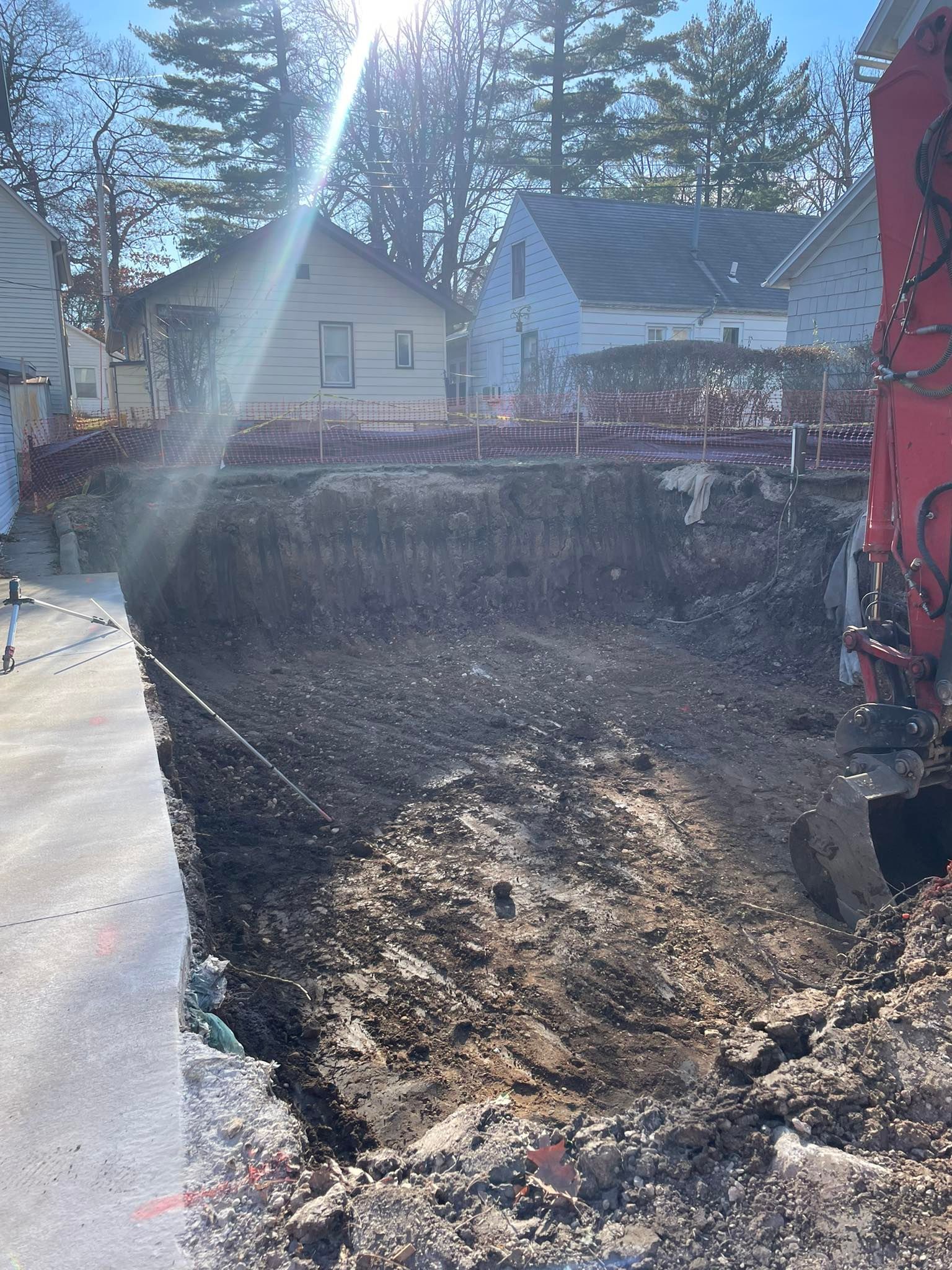 Excavation next to a road, exposing soil layers and adjacent houses on a sunny day.