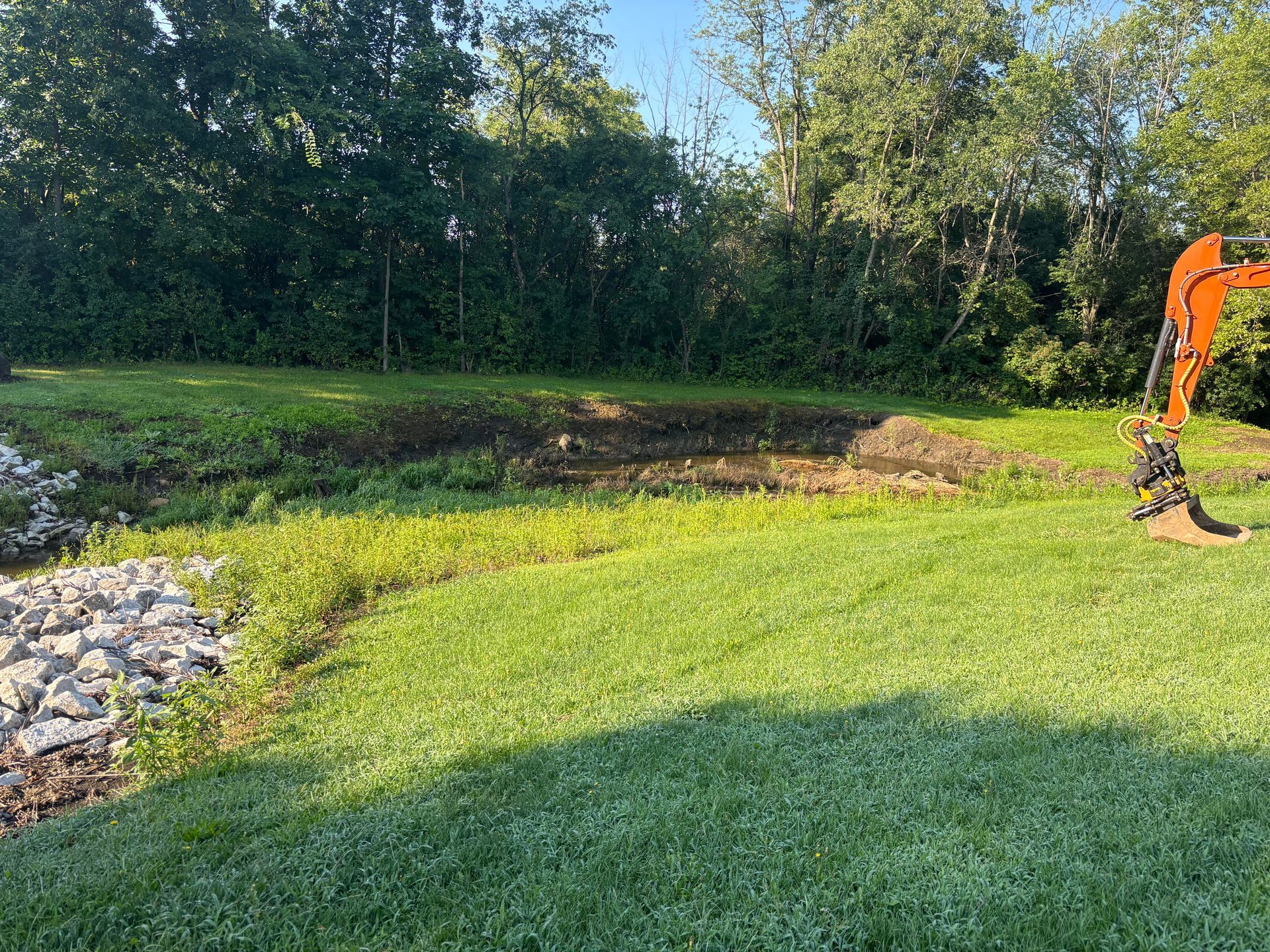 Grassy area with an excavator near a dug-out bank of dirt. Trees are in the background.