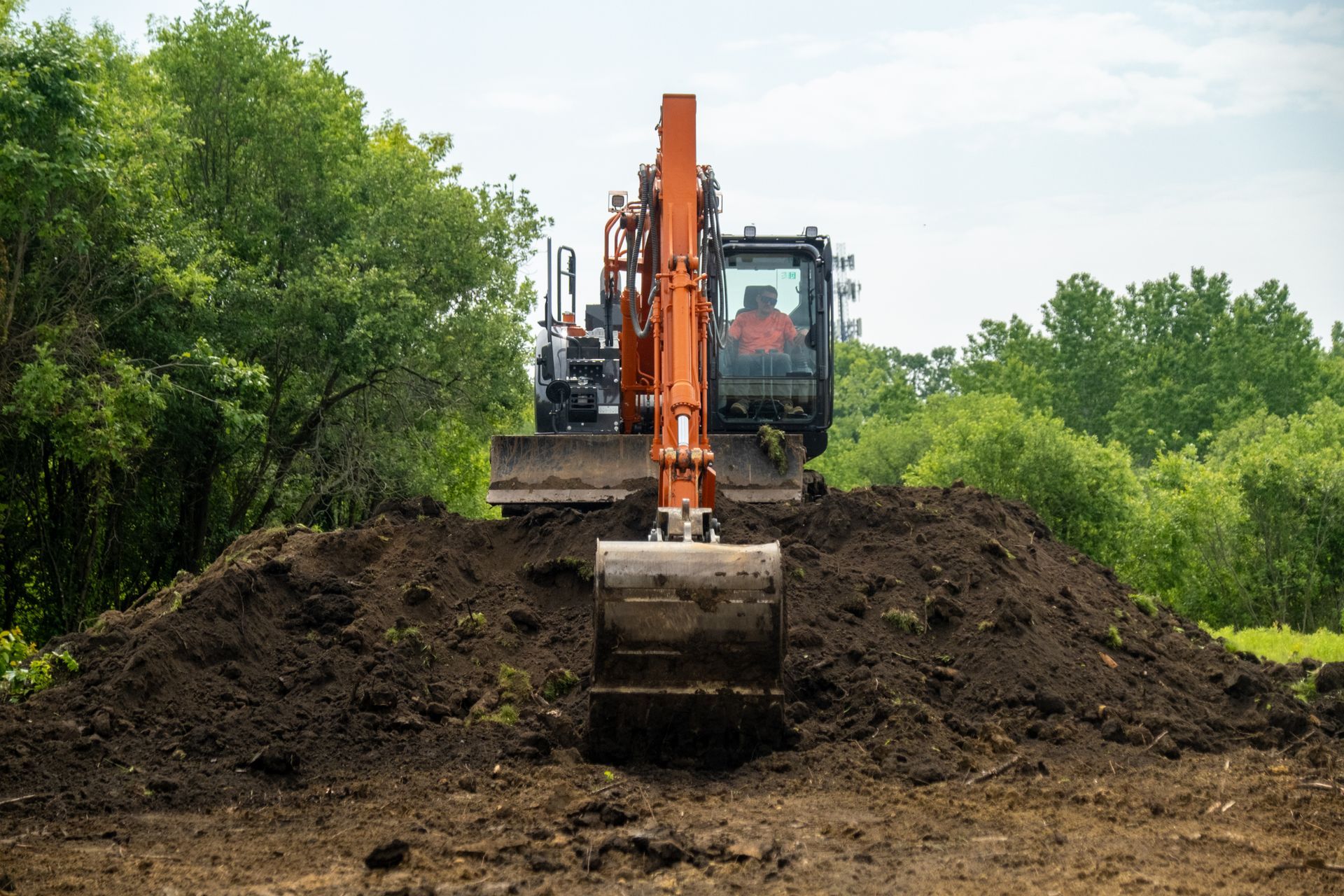 Orange excavator digging dirt, operator in cab, trees in background.