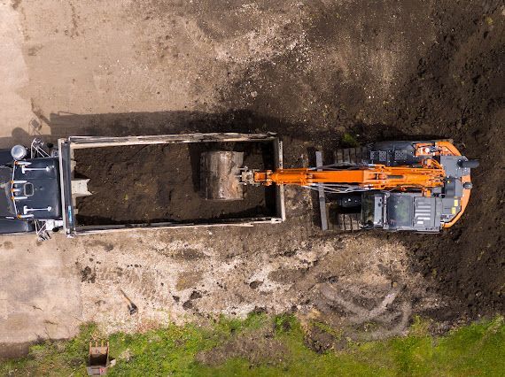 Overhead view of an excavator loading dark soil into a dump truck, outdoors on a concrete surface.