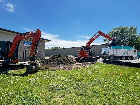 Two excavators loading a dump truck with soil near a building on a sunny day.