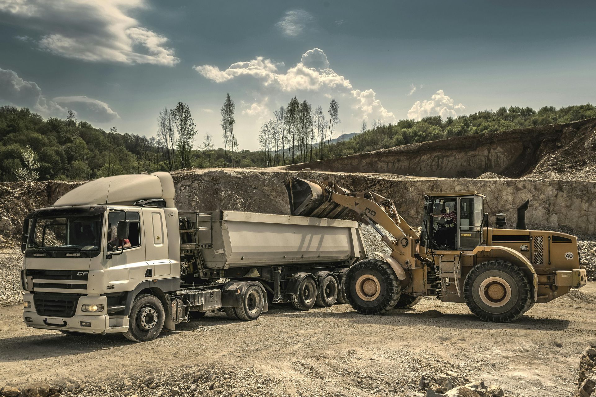 A large, yellow loader filling a dump truck with gravel at a quarry under a cloudy sky.