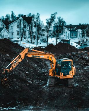 Yellow excavator digging in a dark dirt pile, with snow and buildings in the background.