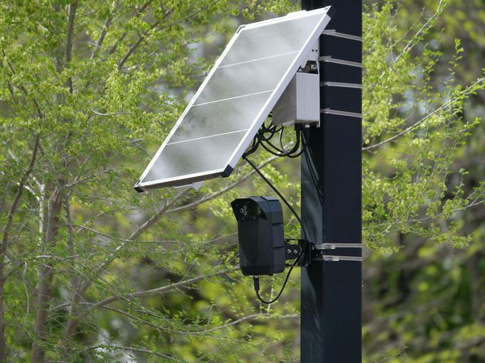 Solar panel mounted on a pole with attached equipment beside leafy trees