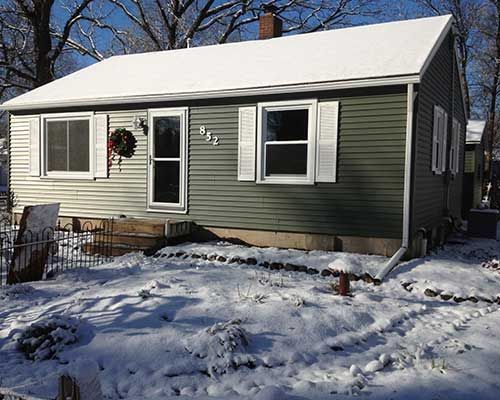 A small house with a green siding and white shutters is covered in snow.